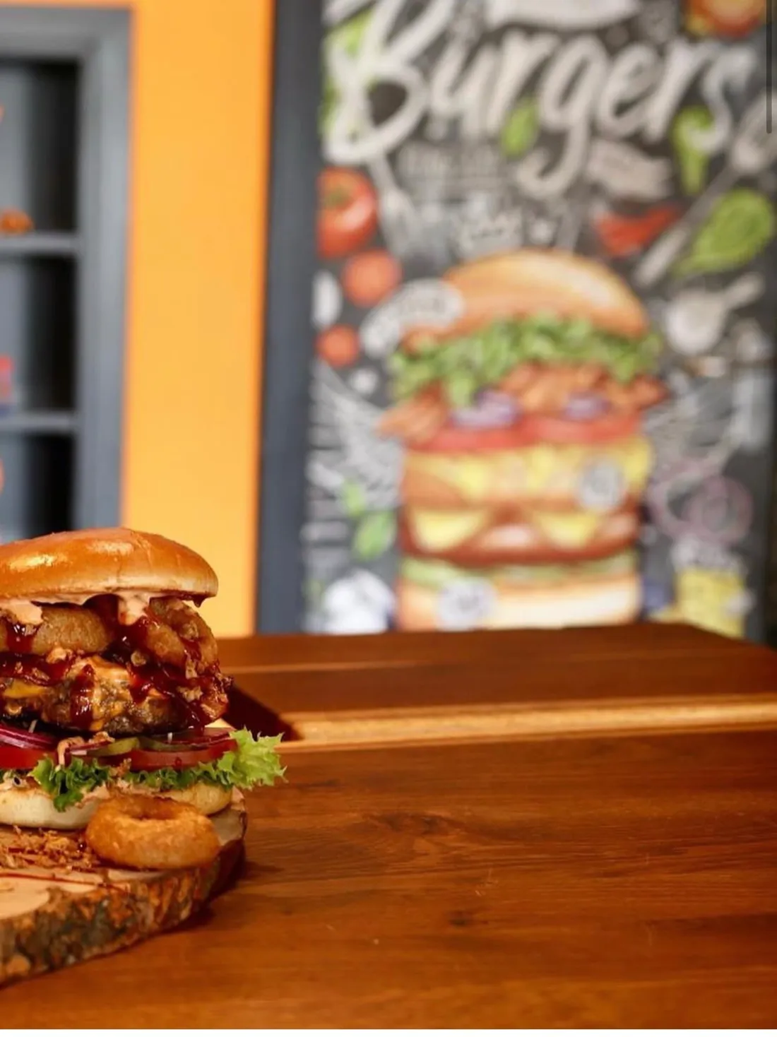 BBQ Burger mit Onion Rings auf einem Holzbrett, im Hintergrund die Kreidetafel im Laden